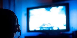 PC Gaming On A TV – A Lazy Gamer’s Guide Young male gamer with glasses and headset playing video game at home in the dark room using game console controller watching at LED TV. Gaming and entertainment concepts