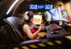 PC Gaming vs. Mobile Gaming: Which is Better? Side profile of Female gamer in red top sitting in black leather chair in front of gaming monitors holding a mobile phone