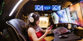 PC Gaming vs. Mobile Gaming: Which is Better? Side profile of Female gamer in red top sitting in black leather chair in front of gaming monitors holding a mobile phone