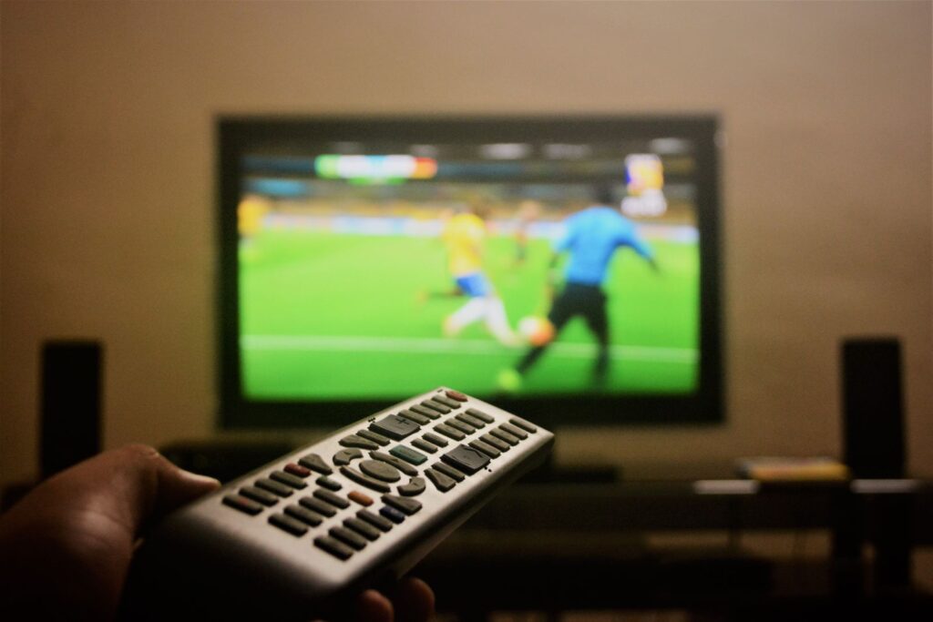 A hand holding a TV remote in front of a television showing a football match