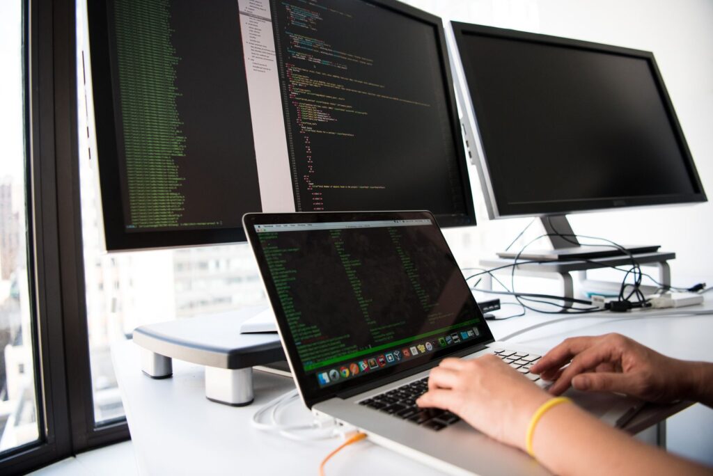 A developer typing on a laptop in front of two large monitors showing lines of code