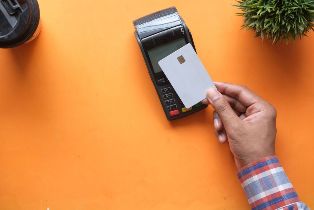 A hand holding a blank payment card above a card terminal on an orange desk