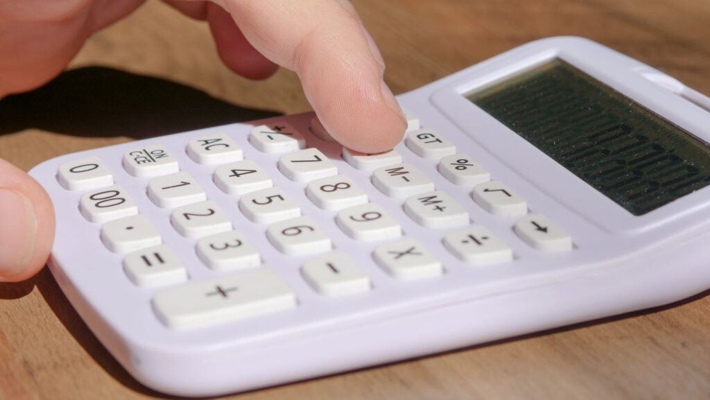 A finger pressing keys on a white calculator on a wooden desk