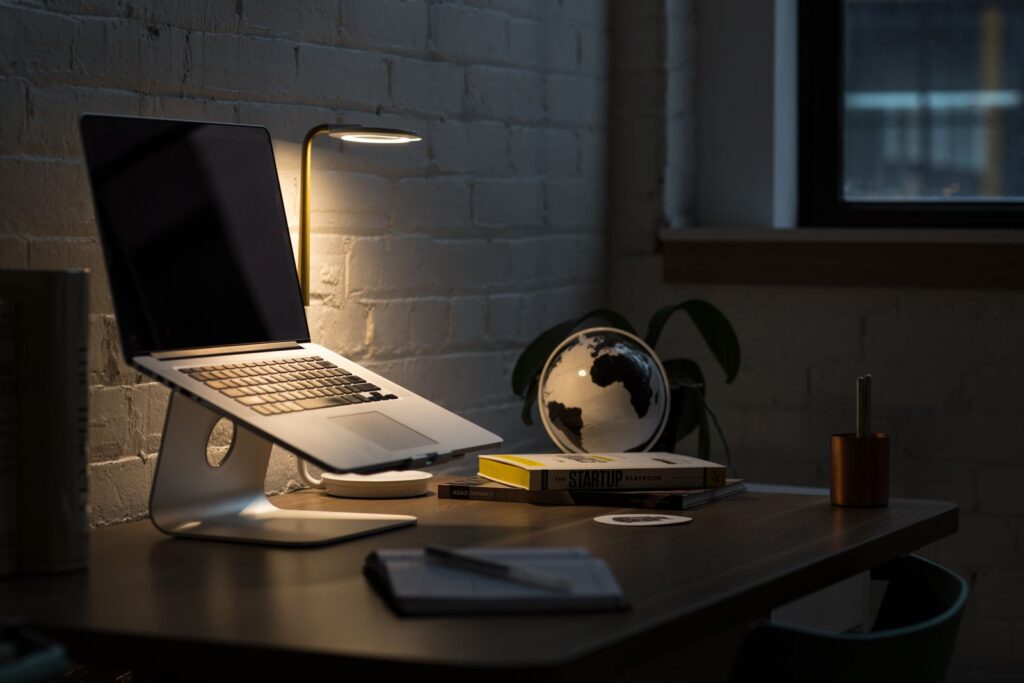 A dimly lit home office desk with a laptop on a stand, a lamp and a small globe