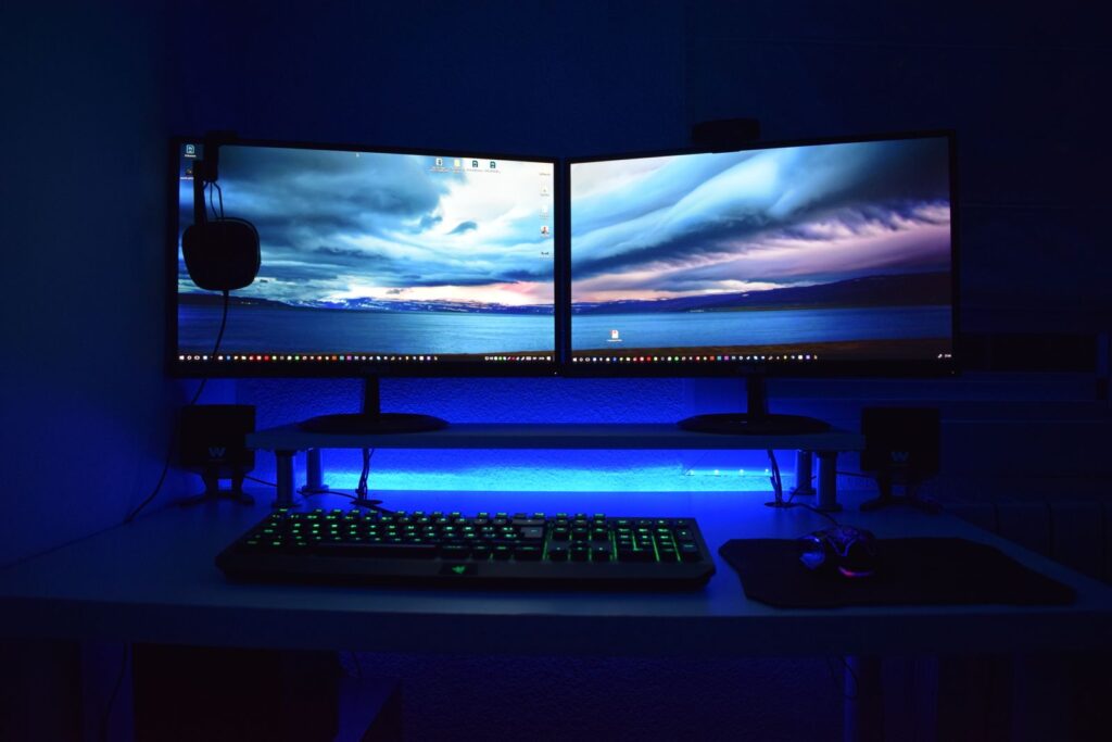 A pair of monitors on a desk lit with blue LED light, with a keyboard in front