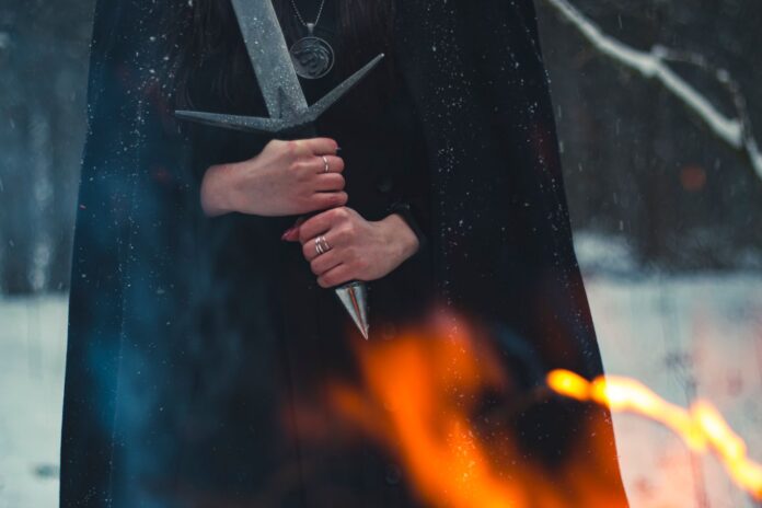 Cloaked figure gripping a sword beside a fire in the snow