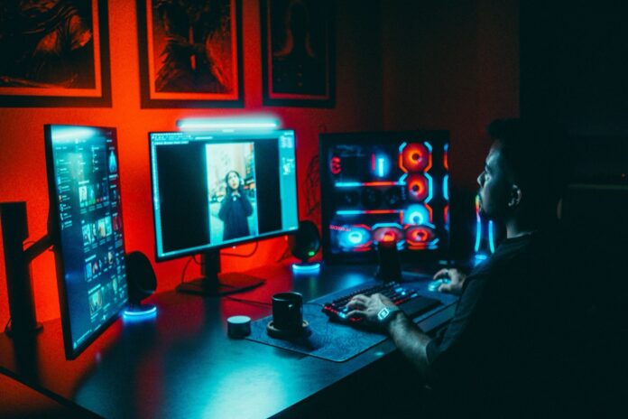 PC desk with three monitors and a glowing custom gaming PC in a dark red lit room